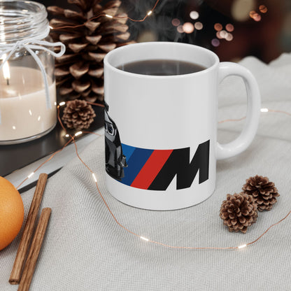 Lifestyle view of the 11oz White Coffee Mug with a BMW M4 GTS with the M emblem placed beside the car.   